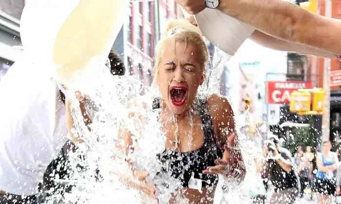 Blonde woman screaming as two people dump buckets of water on her head in an outdoor ice bucket challenge.