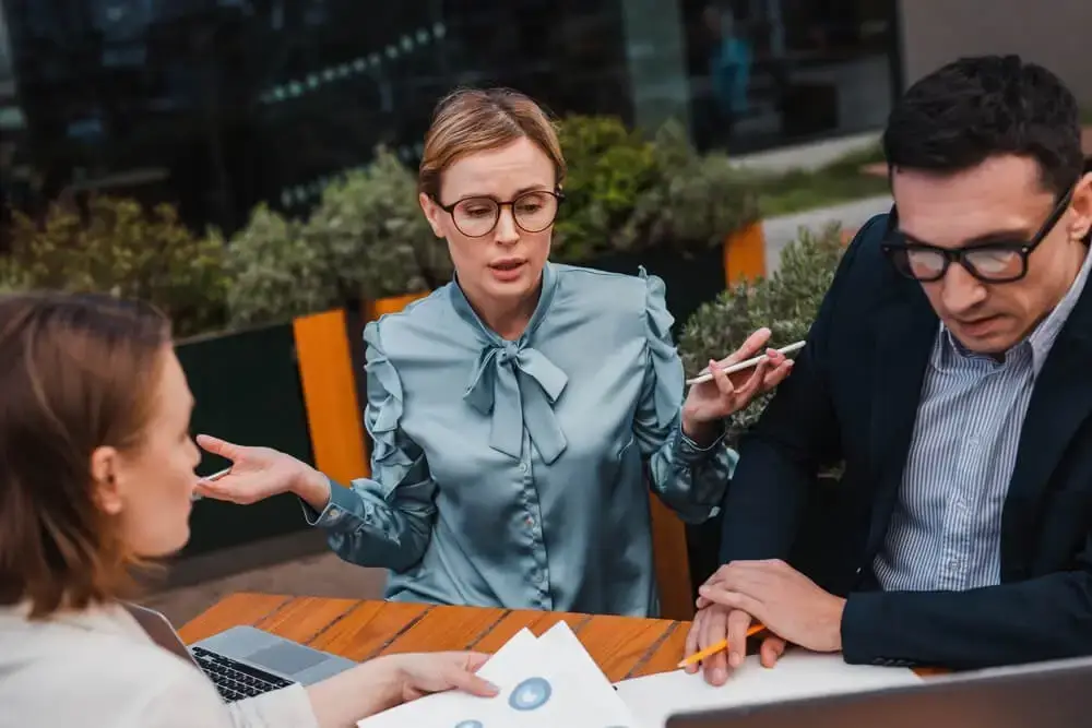 an image of three people sitting around a table with papers and a laptop, engaged in discussion. the group may be discussing various topics related to digital marketing, such as key performance indicators (kpis), strategies, and campaign planning. the scene suggests a collaborative effort among team members, where everyone is contributing their expertise towards achieving shared goals.