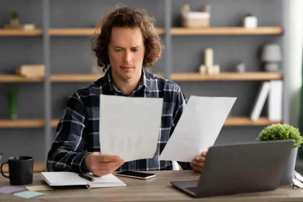Man with curly hair reviewing documents at a desk, laptop and coffee nearby, considering fractional leadership.