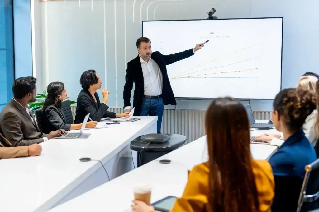 Manager pointing at a sales graph on a large monitor, explaining what CPA in digital marketing means to his diverse team.